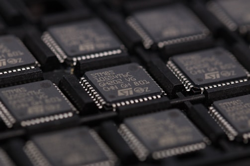 A close-up view of several integrated circuits or microchips arranged in rows. The surface details of the chips are visible, including markings such as part numbers and manufacturer logos. The chips are placed in a dark-colored tray designed for holding them securely.