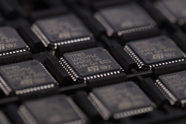 A close-up view of several integrated circuits or microchips arranged in rows. The surface details of the chips are visible, including markings such as part numbers and manufacturer logos. The chips are placed in a dark-colored tray designed for holding them securely.