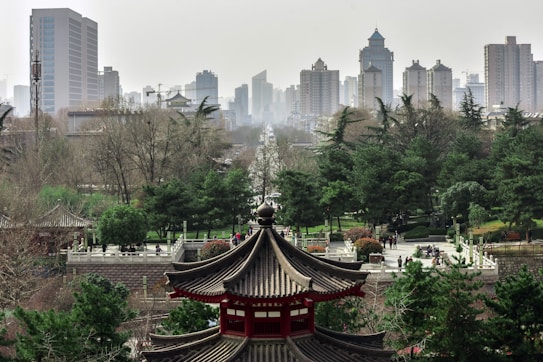An urban park with traditional Chinese architecture in the foreground, and a skyline of modern high-rise buildings in the background. The scene includes lush green trees and a busy park filled with people enjoying the space.