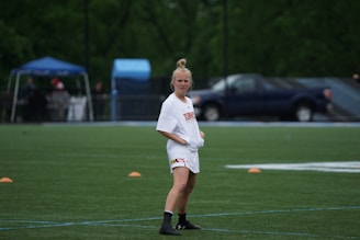 A person wearing a white sports jersey, standing on a green field with orange cones arranged around. A blue tent and a parked truck are visible in the background along with blurred trees.