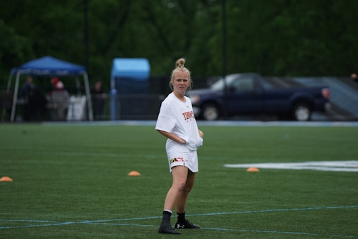 A person wearing a white sports jersey, standing on a green field with orange cones arranged around. A blue tent and a parked truck are visible in the background along with blurred trees.