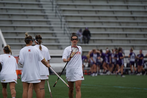 A group of female lacrosse players in white jerseys are gathered on a field. One player is holding a lacrosse stick and wears protective goggles. In the background, a team in purple jerseys is sitting and standing near bleachers.