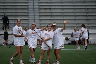 Four women wearing white sports uniforms are standing on a green field with lacrosse sticks. They are smiling and appear to be enjoying themselves. The setting seems to be a stadium with rows of empty bleachers in the background.