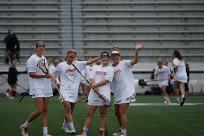 Four women wearing white sports uniforms are standing on a green field with lacrosse sticks. They are smiling and appear to be enjoying themselves. The setting seems to be a stadium with rows of empty bleachers in the background.
