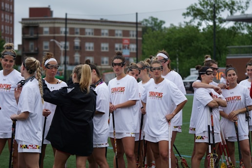 A group of young women dressed in white jerseys congregate on a sports field, some holding lacrosse sticks. They wear protective eyewear, likely preparing for a game or practice. Buildings and greenery are visible in the background.