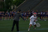 On a sports field, a coach and a player are engaged in what appears to be a practice or warm-up session. The player is dressed in a white uniform and red helmet, holding a lacrosse stick, while the coach is pointing and providing instructions. In the background, a group of players in purple jerseys are lined up, likely for practice or a game.