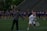 On a sports field, a coach and a player are engaged in what appears to be a practice or warm-up session. The player is dressed in a white uniform and red helmet, holding a lacrosse stick, while the coach is pointing and providing instructions. In the background, a group of players in purple jerseys are lined up, likely for practice or a game.