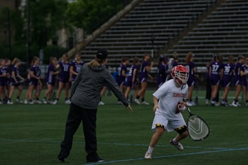 On a sports field, a coach and a player are engaged in what appears to be a practice or warm-up session. The player is dressed in a white uniform and red helmet, holding a lacrosse stick, while the coach is pointing and providing instructions. In the background, a group of players in purple jerseys are lined up, likely for practice or a game.