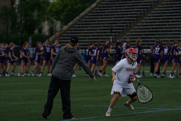 On a sports field, a coach and a player are engaged in what appears to be a practice or warm-up session. The player is dressed in a white uniform and red helmet, holding a lacrosse stick, while the coach is pointing and providing instructions. In the background, a group of players in purple jerseys are lined up, likely for practice or a game.
