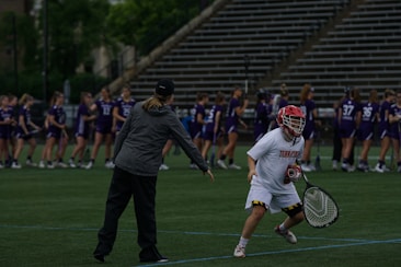On a sports field, a coach and a player are engaged in what appears to be a practice or warm-up session. The player is dressed in a white uniform and red helmet, holding a lacrosse stick, while the coach is pointing and providing instructions. In the background, a group of players in purple jerseys are lined up, likely for practice or a game.