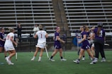 A group of female athletes is playing lacrosse on a sports field. Some players are wearing purple jerseys while others are in white. They are holding lacrosse sticks and wearing protective gear like goggles. The setting is an outdoor stadium with empty bleachers in the background.