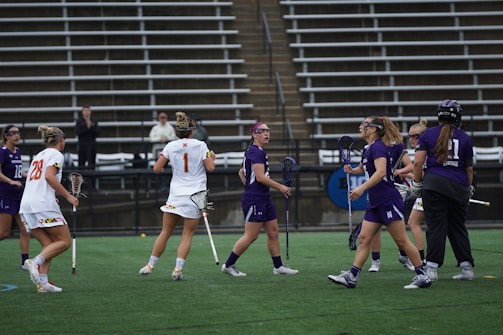 A group of female athletes is playing lacrosse on a sports field. Some players are wearing purple jerseys while others are in white. They are holding lacrosse sticks and wearing protective gear like goggles. The setting is an outdoor stadium with empty bleachers in the background.