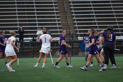 A group of female athletes is playing lacrosse on a sports field. Some players are wearing purple jerseys while others are in white. They are holding lacrosse sticks and wearing protective gear like goggles. The setting is an outdoor stadium with empty bleachers in the background.