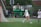 Two athletes in white sports uniforms are on a grassy field, engaging in a game of lacrosse. One player is near the goal, and a net is visible on the left side. A person in the background appears to be observing, with equipment and a green lift in view. The overcast weather suggests a dreary atmosphere.