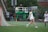 Two athletes in white sports uniforms are on a grassy field, engaging in a game of lacrosse. One player is near the goal, and a net is visible on the left side. A person in the background appears to be observing, with equipment and a green lift in view. The overcast weather suggests a dreary atmosphere.
