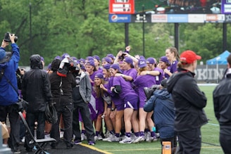 A group of athletes dressed in matching purple uniforms are celebrating on a sports field. They are huddled together, smiling and raising their arms in excitement. Several photographers and cameramen in rain gear are capturing the moment, indicating that the event is taking place outdoors in rainy weather. The background includes a scoreboard and lush green trees.
