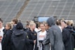 A group of people, likely athletes, are gathered together on a sports field. They are wearing black jackets with some individuals dressed in white sports uniforms underneath. A few people have their hair tied up in buns, and most are engaged in conversation. The background shows tiered stadium seats, suggesting the setting is an outdoor sports venue.