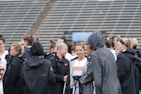 A group of people, likely athletes, are gathered together on a sports field. They are wearing black jackets with some individuals dressed in white sports uniforms underneath. A few people have their hair tied up in buns, and most are engaged in conversation. The background shows tiered stadium seats, suggesting the setting is an outdoor sports venue.