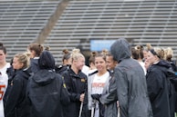 A group of people, likely athletes, are gathered together on a sports field. They are wearing black jackets with some individuals dressed in white sports uniforms underneath. A few people have their hair tied up in buns, and most are engaged in conversation. The background shows tiered stadium seats, suggesting the setting is an outdoor sports venue.