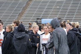 A group of people, likely athletes, are gathered together on a sports field. They are wearing black jackets with some individuals dressed in white sports uniforms underneath. A few people have their hair tied up in buns, and most are engaged in conversation. The background shows tiered stadium seats, suggesting the setting is an outdoor sports venue.