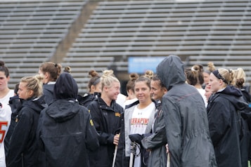 A group of people, likely athletes, are gathered together on a sports field. They are wearing black jackets with some individuals dressed in white sports uniforms underneath. A few people have their hair tied up in buns, and most are engaged in conversation. The background shows tiered stadium seats, suggesting the setting is an outdoor sports venue.