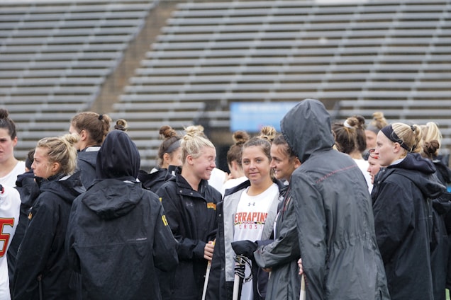 A group of people, likely athletes, are gathered together on a sports field. They are wearing black jackets with some individuals dressed in white sports uniforms underneath. A few people have their hair tied up in buns, and most are engaged in conversation. The background shows tiered stadium seats, suggesting the setting is an outdoor sports venue.