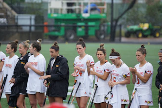 A group of female athletes is standing in a line on a sports field. They are wearing white sports uniforms with 'Maryland' emblazoned on them, and some are holding lacrosse sticks. The background shows a blurred image of a field and some equipment.