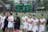 A group of female athletes is standing in a line on a sports field. They are wearing white sports uniforms with 'Maryland' emblazoned on them, and some are holding lacrosse sticks. The background shows a blurred image of a field and some equipment.
