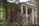 A large, two-story brick house is surrounded by lush greenery. The architecture features tall white columns supporting a front porch with a balcony. There are several windows visible, and the facade is partially obscured by various trees and shrubs.