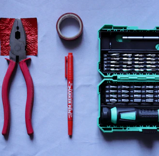A collection of tools placed on a flat surface includes red-handled pliers, a roll of red tape, a red marker pen, and a green tool case filled with various screwdriver bits and a handle.