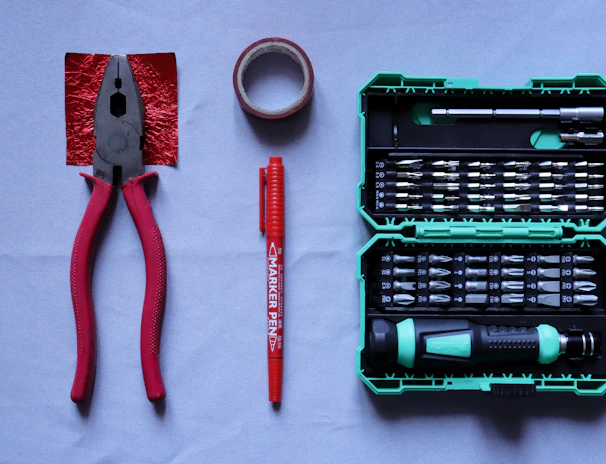 A collection of tools placed on a flat surface includes red-handled pliers, a roll of red tape, a red marker pen, and a green tool case filled with various screwdriver bits and a handle.