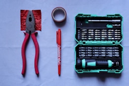 A collection of tools placed on a flat surface includes red-handled pliers, a roll of red tape, a red marker pen, and a green tool case filled with various screwdriver bits and a handle.