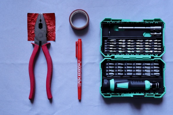 A collection of tools placed on a flat surface includes red-handled pliers, a roll of red tape, a red marker pen, and a green tool case filled with various screwdriver bits and a handle.