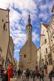 A bustling street scene in a historic European city features a tall stone tower centered between classic architectural buildings. People in winter clothing are walking through the street, engaging in various activities under a partly cloudy sky.