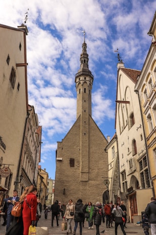 A bustling street scene in a historic European city features a tall stone tower centered between classic architectural buildings. People in winter clothing are walking through the street, engaging in various activities under a partly cloudy sky.