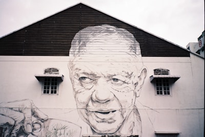 A large mural is painted on the side of a building, depicting the face of an elderly man with detailed features. He appears to be holding a cup, adding realism to the artwork. The mural covers the upper floors of a structure with windows and ventilation elements.