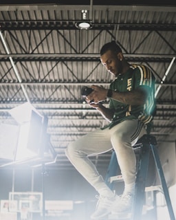 A person sitting on a ladder indoors, examining a camera, with bright studio lighting illuminating the area.
