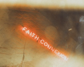 A blurred photograph featuring a neon sign that reads 'FAITH CONNEXION' with a warm, diffused lighting effect across the image. The background is abstract with a vintage, textured appearance.