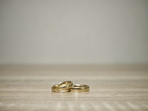 Two gold rings are placed on a light wooden surface with a neutral gray background, creating a minimalist and elegant composition.