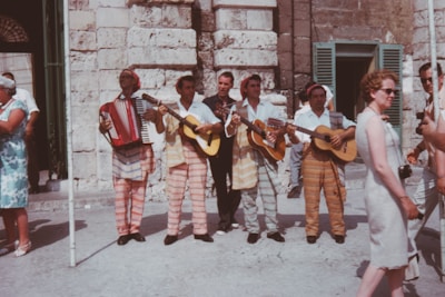 A group of musicians dressed in striped clothing are playing instruments, including guitars and an accordion, in front of a stone building. Several people are standing and walking nearby, with one woman in a light dress and sunglasses carrying a camera. The scene appears to be lively and outdoors.
