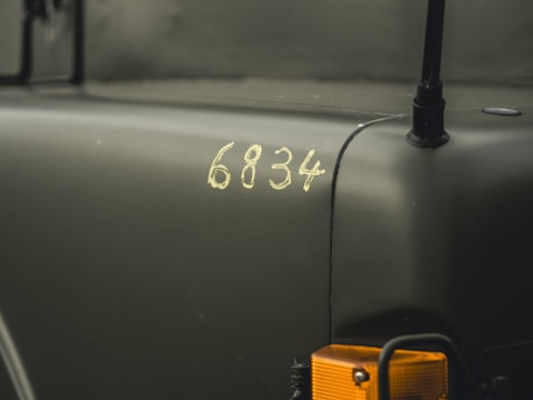 A close-up view of a military vehicle with the number 6834 displayed on a matte dark green surface. The image includes an orange indicator light and parts of the vehicle's structure.