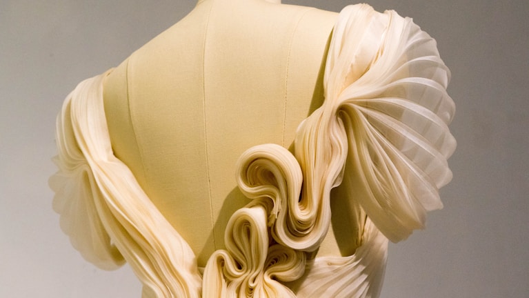 An intricately designed garment with pleated, flowing fabric in a soft cream color, featuring elaborate folds and ruffles that create a sculptural look. The fabric appears delicate and is arranged in an artistic, almost organic form, highlighting the elegance of the design.