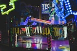 Brightly lit amusement ride with vibrant, multi-colored lights illuminating the surroundings. The ride features chairs and a structural arm, situated indoors with themed decorations in the background, suggestive of a festive or fair-like atmosphere.