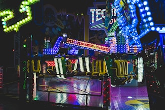 Brightly lit amusement ride with vibrant, multi-colored lights illuminating the surroundings. The ride features chairs and a structural arm, situated indoors with themed decorations in the background, suggestive of a festive or fair-like atmosphere.