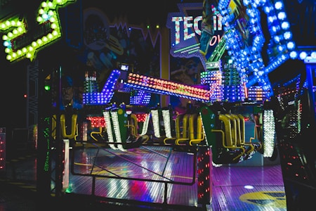 Brightly lit amusement ride with vibrant, multi-colored lights illuminating the surroundings. The ride features chairs and a structural arm, situated indoors with themed decorations in the background, suggestive of a festive or fair-like atmosphere.