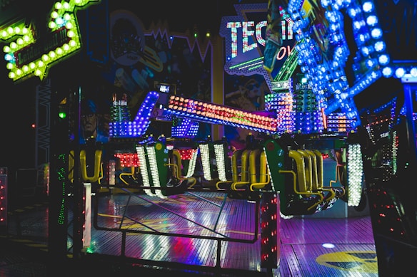 Brightly lit amusement ride with vibrant, multi-colored lights illuminating the surroundings. The ride features chairs and a structural arm, situated indoors with themed decorations in the background, suggestive of a festive or fair-like atmosphere.