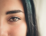 A close-up of a human eye with long eyelashes and a well-defined eyebrow. The skin is smooth, and there is a subtle presence of blush or natural redness on the cheek area.