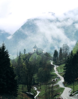 A serene mountain landscape with a winding path leading through lush, green trees and a small stream flowing alongside. In the distance, a quaint building with a pointed roof sits atop a hill. Mist and clouds hover over the scene, partially obscuring the mountain peaks and adding an ethereal feel.