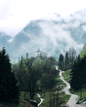 A serene mountain landscape with a winding path leading through lush, green trees and a small stream flowing alongside. In the distance, a quaint building with a pointed roof sits atop a hill. Mist and clouds hover over the scene, partially obscuring the mountain peaks and adding an ethereal feel.