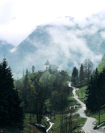 A serene mountain landscape with a winding path leading through lush, green trees and a small stream flowing alongside. In the distance, a quaint building with a pointed roof sits atop a hill. Mist and clouds hover over the scene, partially obscuring the mountain peaks and adding an ethereal feel.